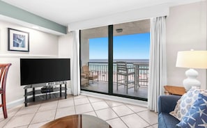 A hotel room with a TV, a balcony with a view of the beach, and a sofa with a starfish pillow.