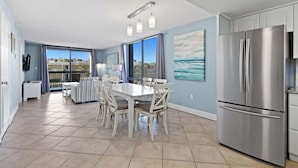 A modern kitchen with a large stainless steel refrigerator, a dining area with white chairs and a table, and a balcony with a view of the ocean. - SunDestin 2BR Beach Condo With Modern Updates and Spacious Layout - Unit 0218 (Destin)
