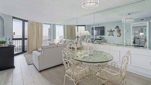 Uma sala de estar moderna com uma mesa de jantar de vidro, sofás brancos e um espelho grande refletindo o ambiente.