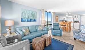 A modern living room with a blue sofa, a grey armchair, and a patterned armchair. There is a dining area with wooden furniture and a kitchen in the background. - SunDestin High-Floor 2BR Condo With Gulf Views and Private Balcony (Destin)