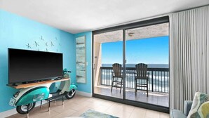 A room with a television, teal scooter, and a balcony with chairs and a view of the ocean.