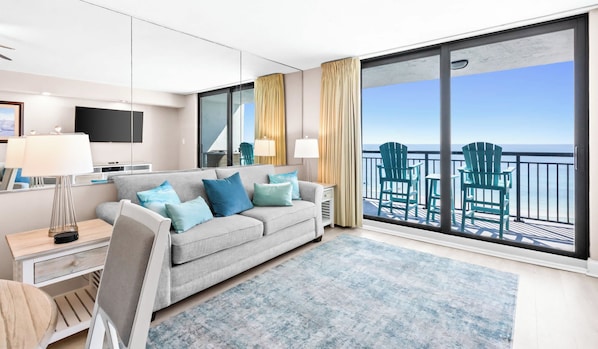 A modern hotel room with a large balcony, a sofa with cushions, a TV, and a desk with a lamp.