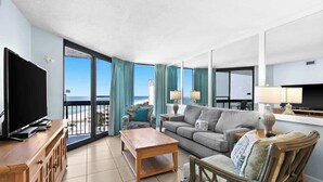 Ein geräumiges Wohnzimmer mit einem großen Fernseher, einem Holztisch, einer grauen Couch und einem Korbstuhl. Es gibt einen Balkon mit Blick auf den Ozean und ein Gebäude im Hintergrund.