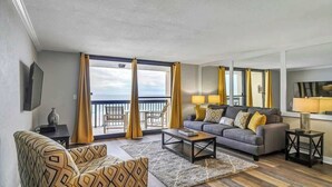 A modern living room with a grey sofa, patterned armchairs, a coffee table, and a TV. Large windows with yellow curtains offer a view of the balcony. - SunDestin Beach Retreat With Bar Seating and Sunset Views - Unit 0508 (Destin)