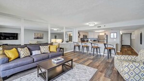 A modern living room with a grey sofa, coffee table, and patterned cushions. A kitchen area with a bar stool and a dining table is visible in the background. - SunDestin Beach Retreat With Bar Seating and Sunset Views - Unit 0508 (Destin)