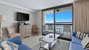A modern living room with a flat-screen TV, a glass coffee table, and a sofa. There is a balcony with a view of the ocean. - SunDestin Resort Unit 1510 (Destin)