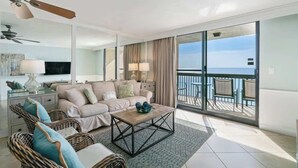 Amplia sala de estar con un sofá grande, una mesa de centro y sillas de mimbre. Hay un balcón con vista al mar.