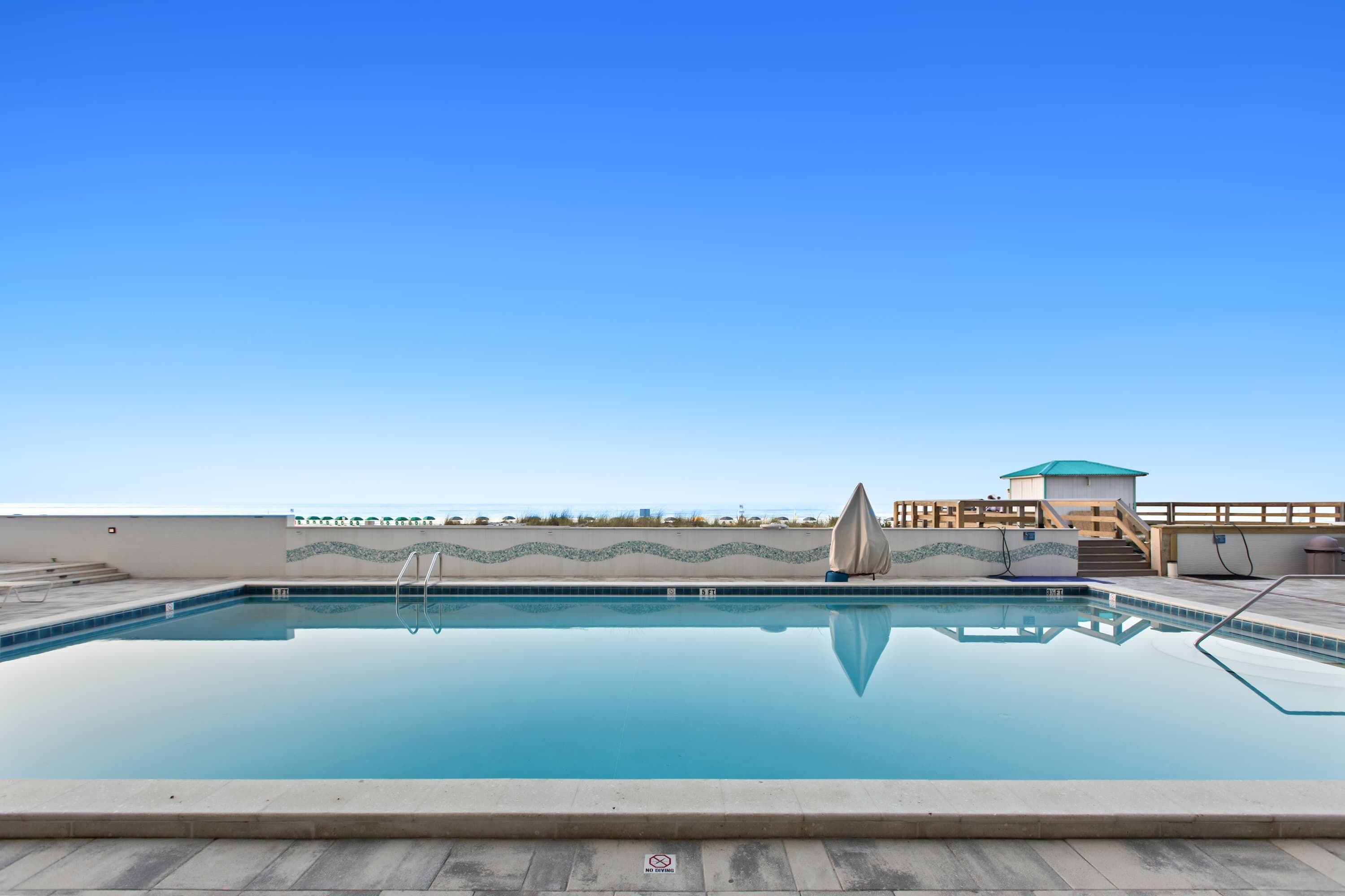 Piscina retangular com céu azul claro ao fundo, uma torre de salva-vidas e uma área coberta com degraus que levam à piscina.