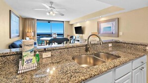 A hotel room with a granite countertop, a sink, a ceiling fan, a large window with a view of the ocean, a flat-screen TV, and a dining area with a table and chairs. - Beachfront condo in Destin with balcony, pool , and resort amenities (Destin)