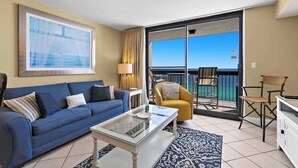 A living room with a blue sofa, a glass coffee table, and a yellow armchair. There is a large window with a view of the ocean. - Beachfront condo in Destin with balcony, pool , and resort amenities (Destin)
