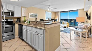 A modern kitchen with white cabinets, a granite countertop, and stainless steel appliances. There is a dining area with a table and chairs, and a large window offering a view. - Beachfront condo in Destin with balcony, pool , and resort amenities (Destin)