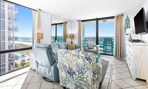 A modern hotel room with a balcony, a sofa, a chair, a TV, and a view of the beach.