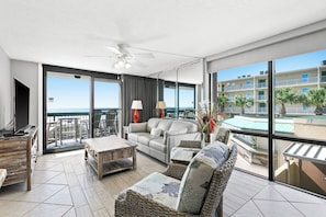 A spacious living area with a balcony, a ceiling fan, and a view of a pool and buildings.