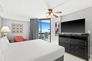 A hotel room with a bed, a television, a ceiling fan, a balcony with a view, and a desk with drawers.