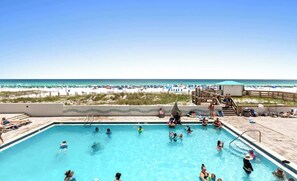 A swimming pool with people swimming and lounging, a beach with umbrellas and people in the background, and a wooden deck with chairs and a lifeguard stand. - SunDestin Resort Unit 0204 (Destin)