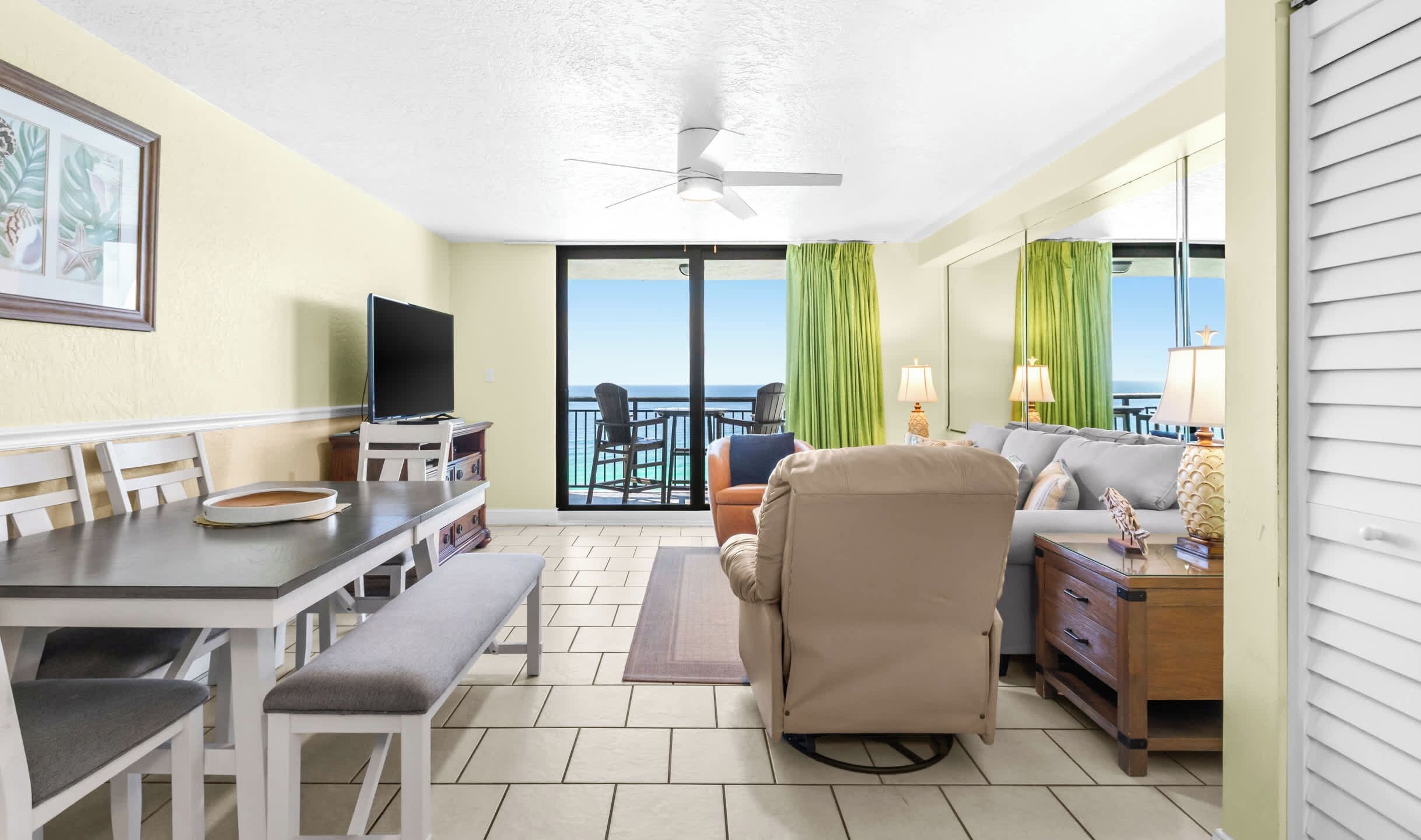 A hotel room with a balcony, a dining area, a sofa, a TV, and a view of the ocean.