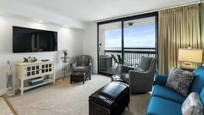 Een moderne woonkamer met een grote televisie, een blauwe bank, een grijze fauteuil, een witte kast en een balkon met uitzicht.