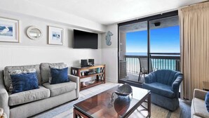 A living room with a grey sofa, a blue armchair, a coffee table, and a flat-screen TV. There is a large window with a view of the ocean. - SunDestin Resort Unit 0710 (Destin)