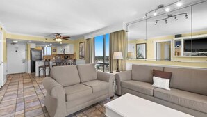 A spacious living area with a sofa, a coffee table, and a TV. There is a kitchen in the background with a dining area. - Spacious 2BR Destin Condo With Gulf Views and Private Balcony (Destin)