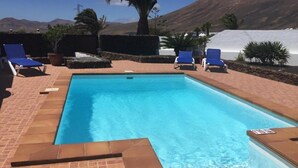 A rectangular swimming pool with blue water, surrounded by a brick patio. There are blue lounge chairs and potted plants around the pool. - Villa Lucky (Tías)