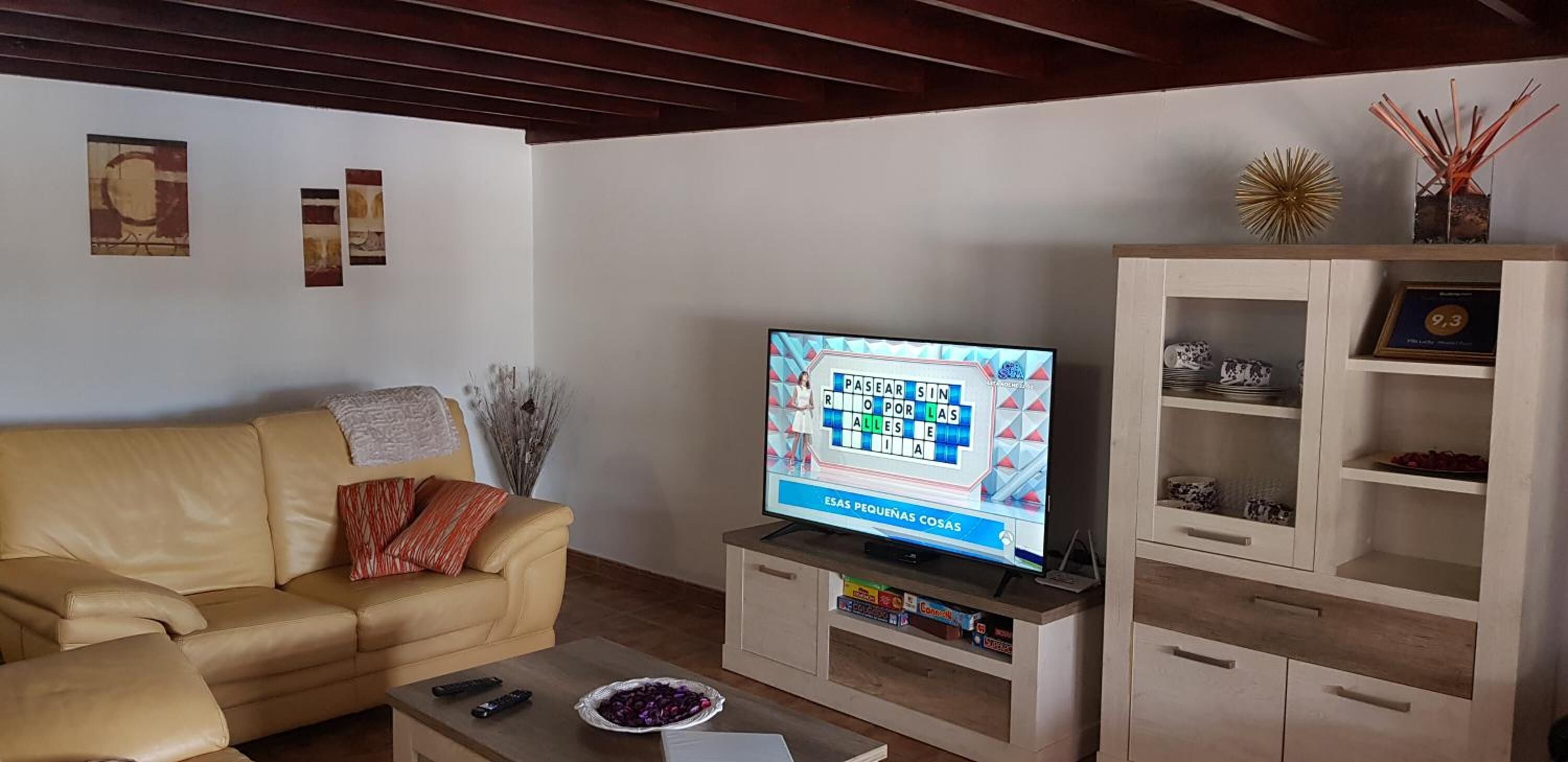 Een woonkamer met een beige bank, een bijzettafeltje met een fruitschaal, een tv waarop een spel wordt vertoond, en een kast met decoratieve objecten.