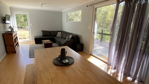 A spacious living room with a wooden dining table, a black leather sofa, and a sliding glass door leading to a deck. - A beautiful chalet in a secluded bush environment, only a short walk to town. (Denmark)