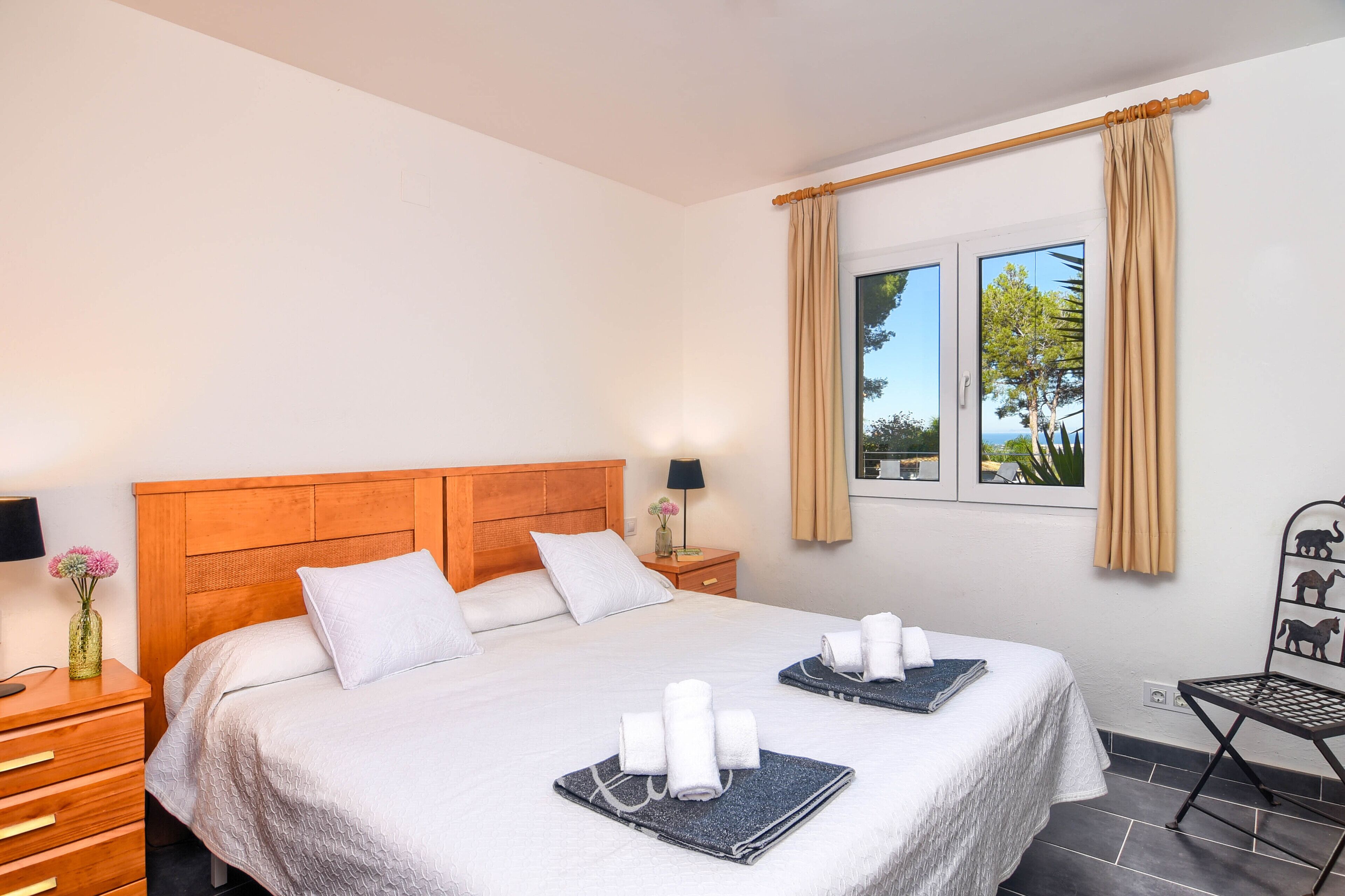 A hotel room with a large bed, wooden headboard, white bedding, a nightstand with a lamp, a window with curtains, and a small table with towels.