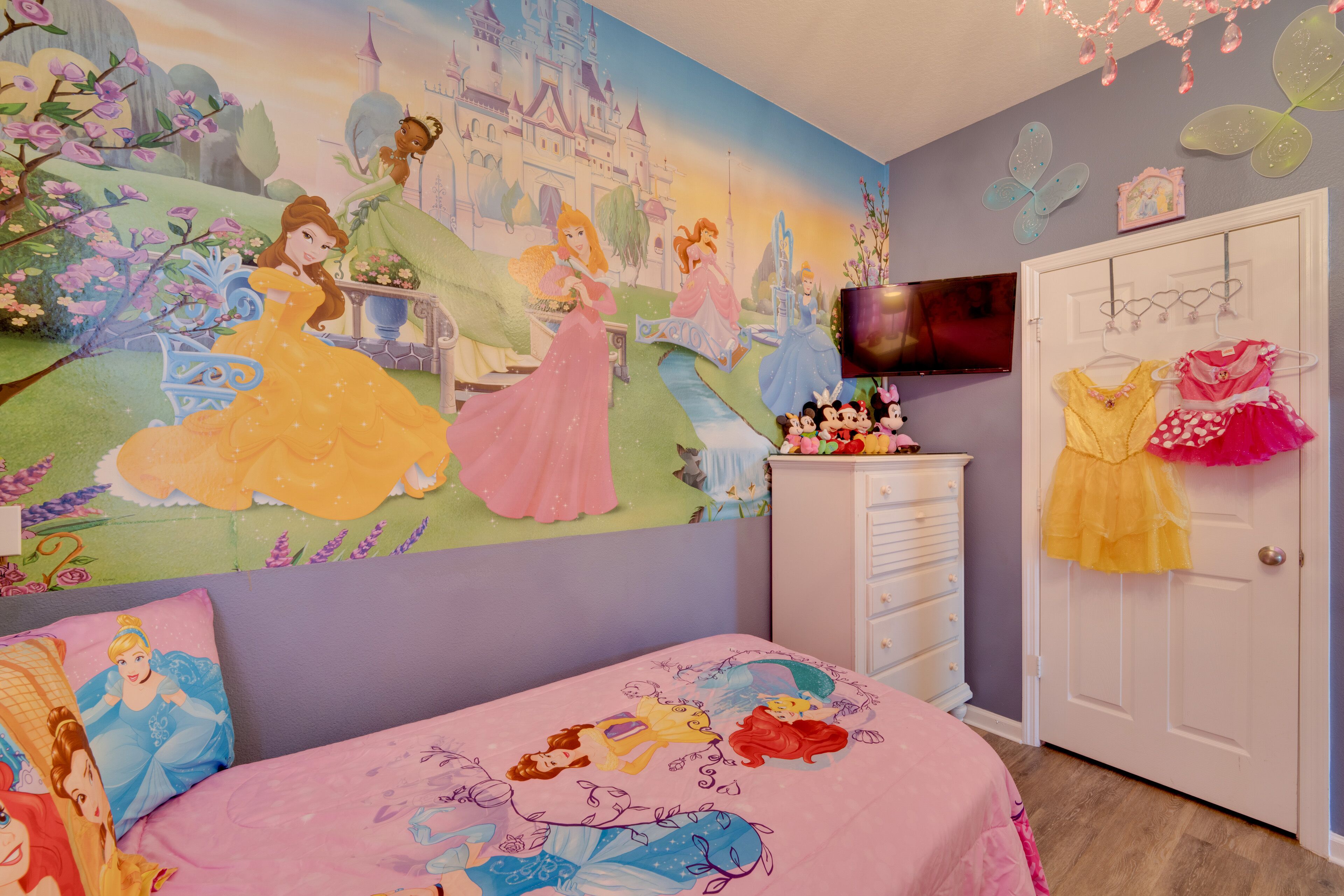 Kinderzimmer mit Wandgemälde von Disney-Prinzessinnen, rosa Bettdecke und Kommode mit Fernseher.