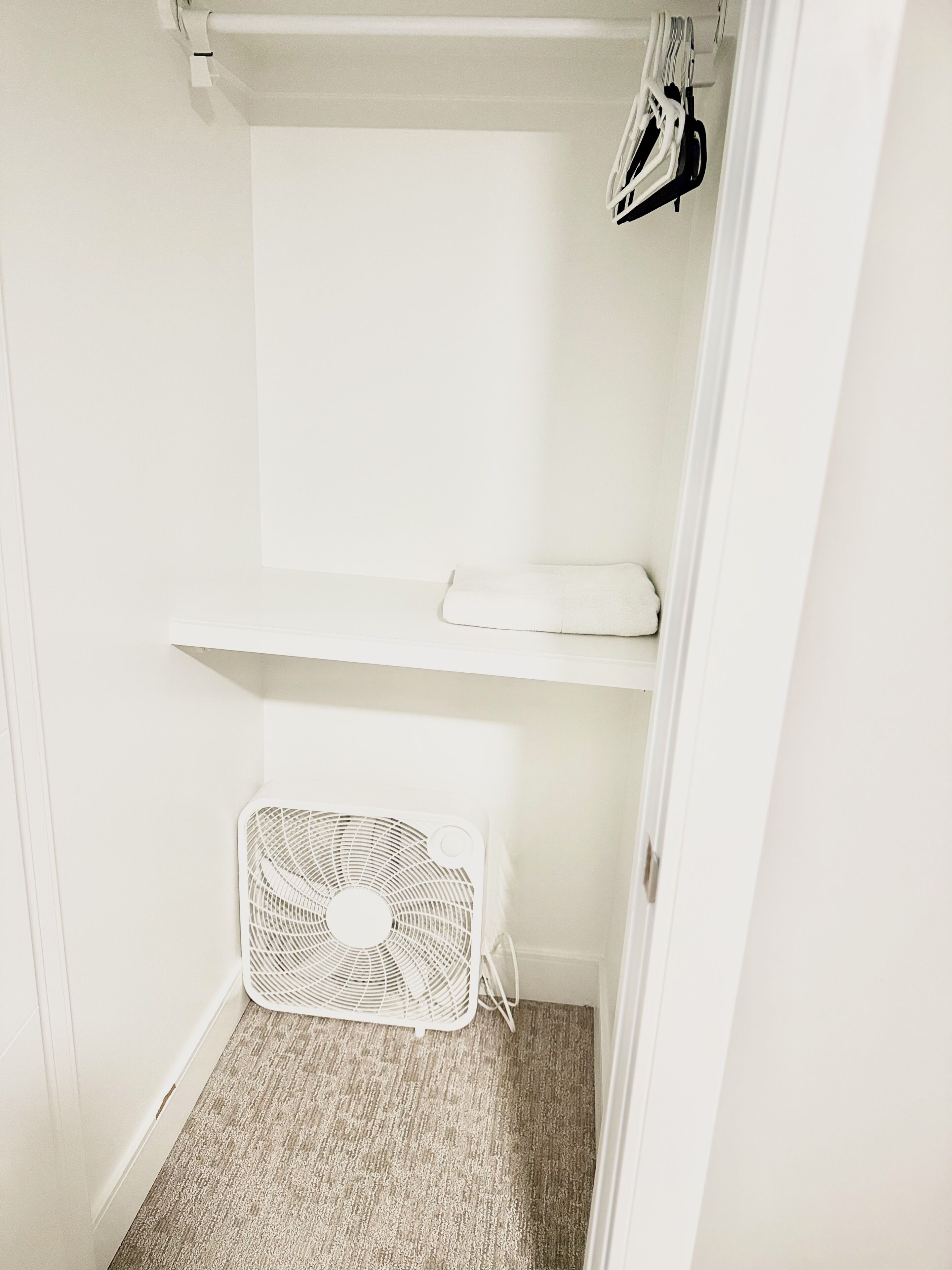 A white closet with a shelf, a hanging rack, and a white fan on the floor.