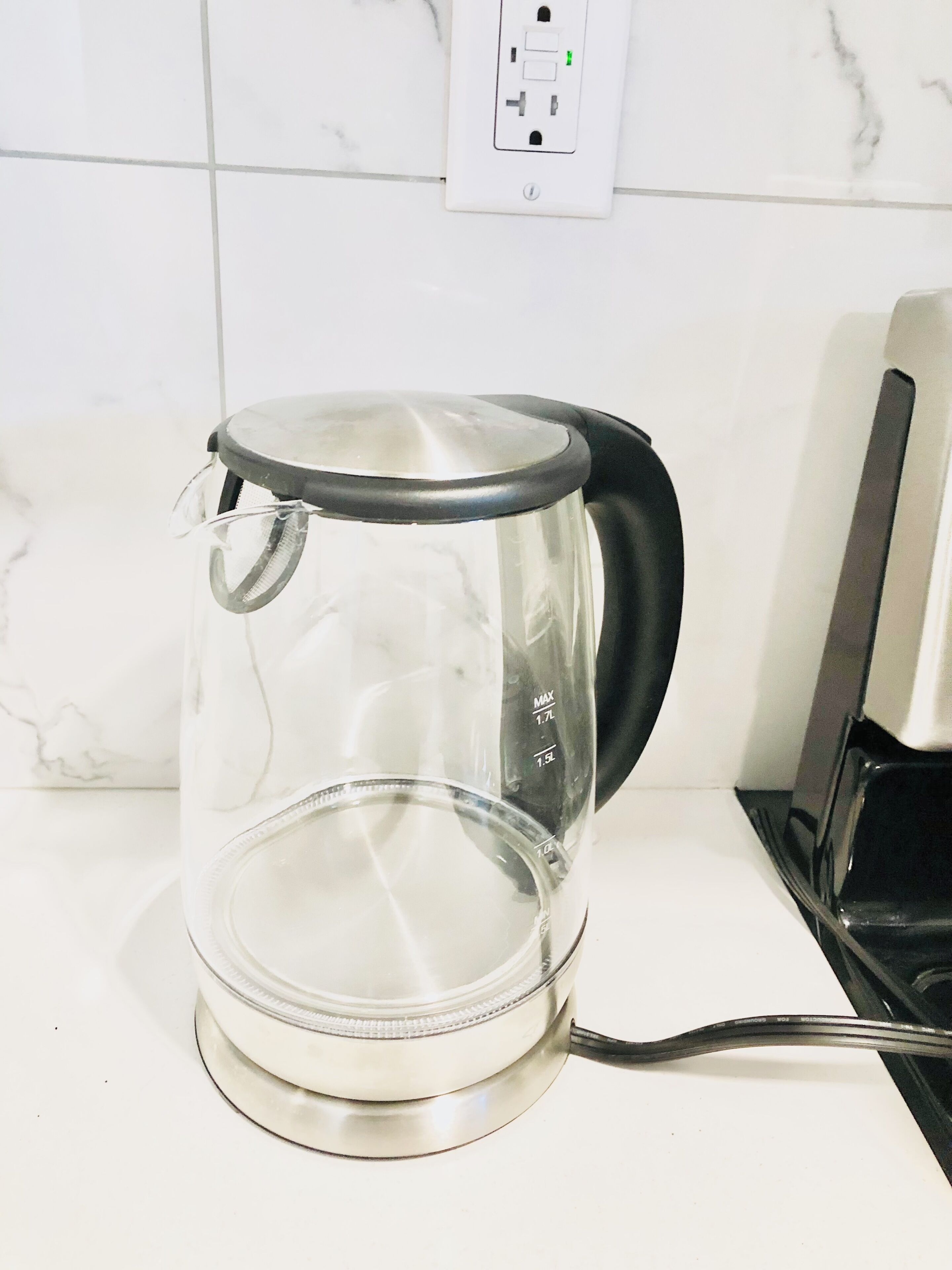 Kirkas lasinen sähkökeitin, jossa on musta kahva ja joka on sijoitettu valkoiselle työtasolle.