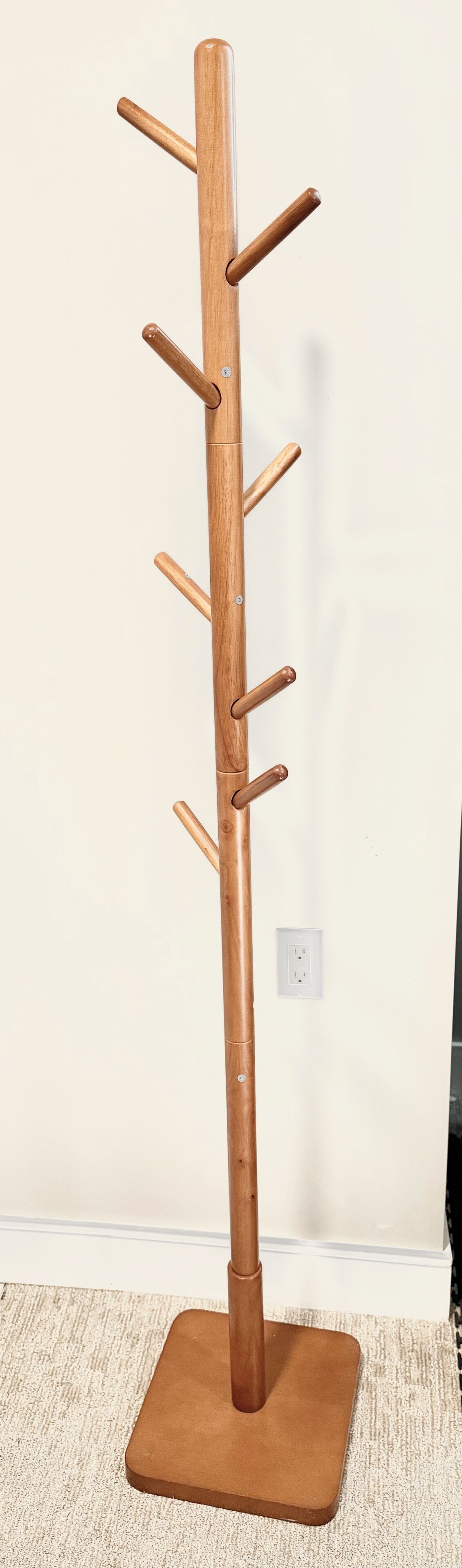 A tall wooden coat rack with multiple hooks, a square base, and an electrical outlet on the wall.