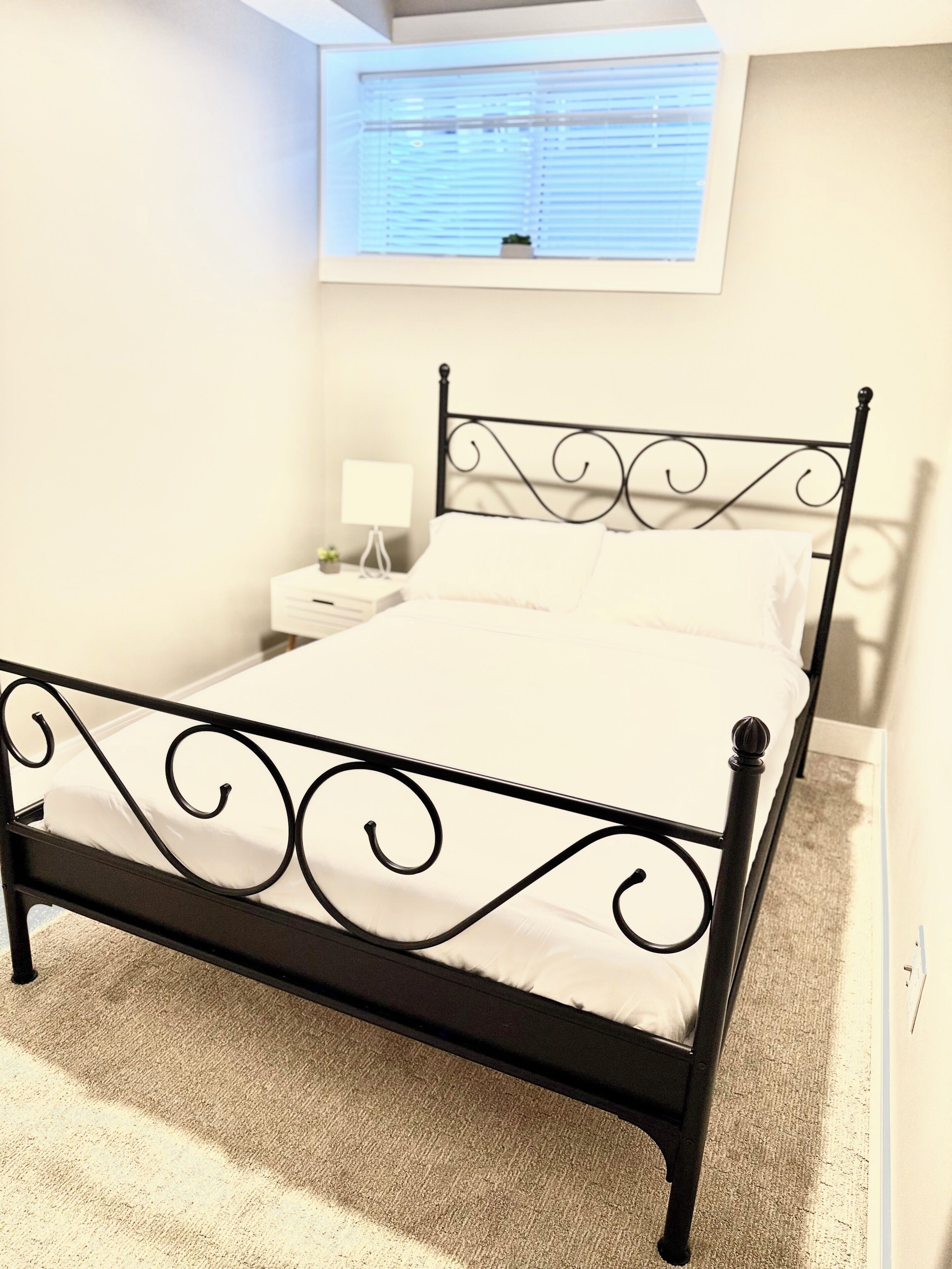 Una cama con estructura metálica, una mesita de noche con lámpara y una ventana con persianas.