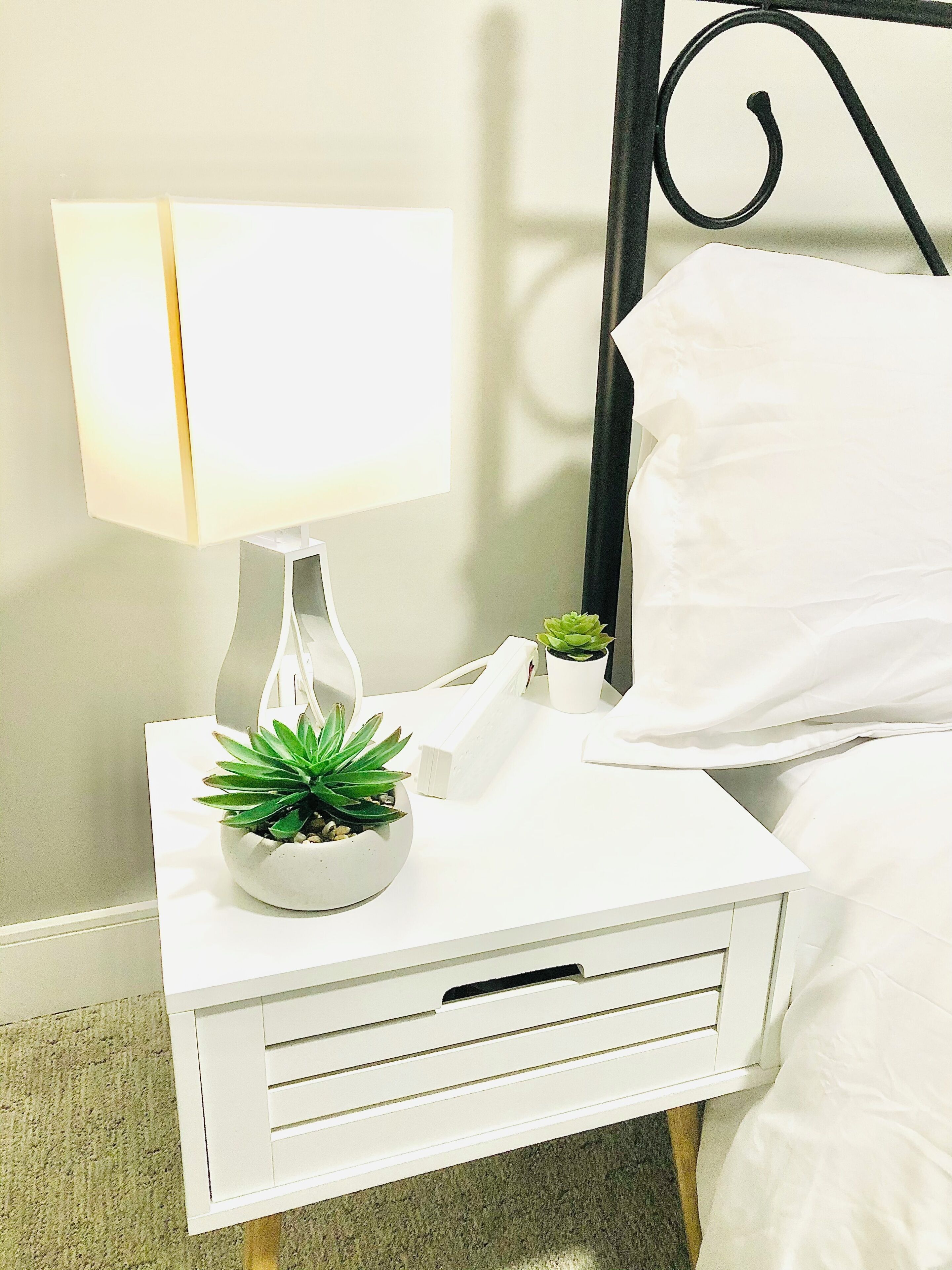 A neatly made bed with white linens, a bedside lamp, a small potted plant, and a white nightstand with a drawer.