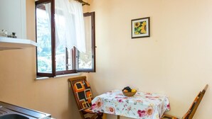 A small dining area with a table covered by a floral tablecloth, two chairs, a window with curtains, and a framed picture on the wall. - Apartments Neve (Dubrovnik)