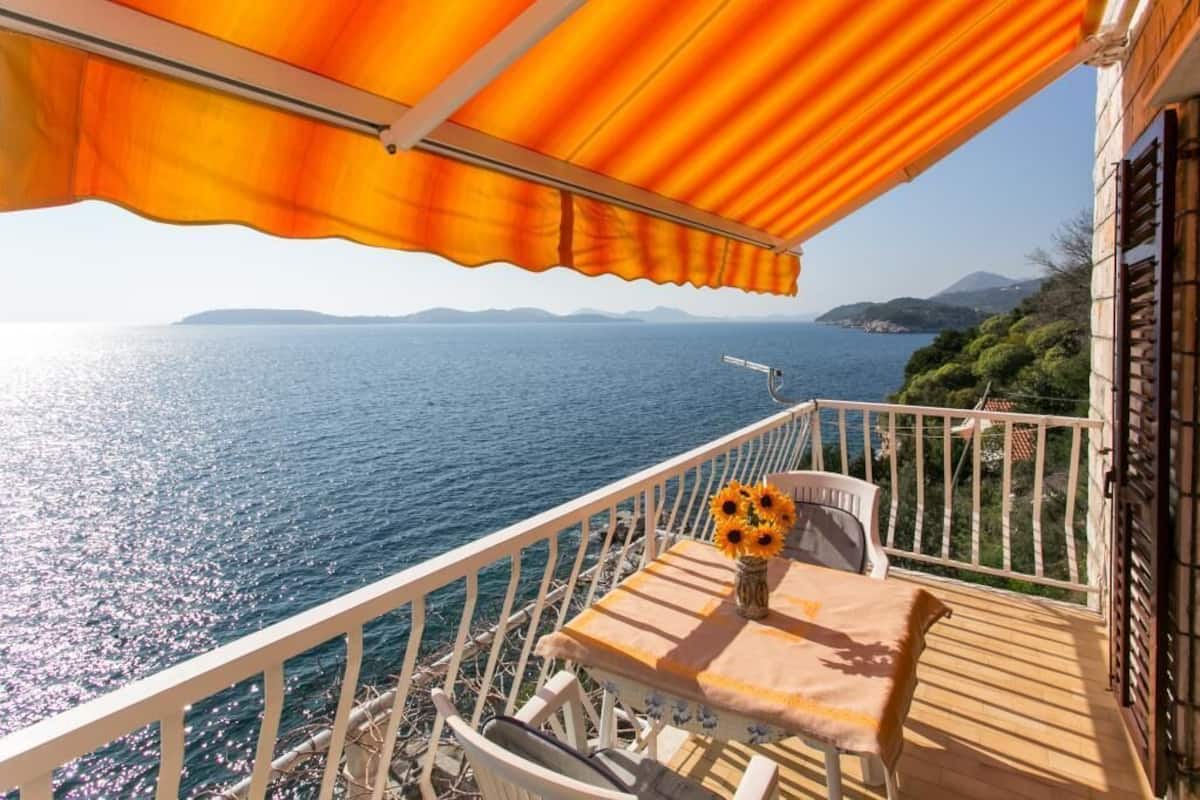 Uma varanda com mesa e cadeiras, um vaso de girassóis e vista para o mar e colinas ao longe.