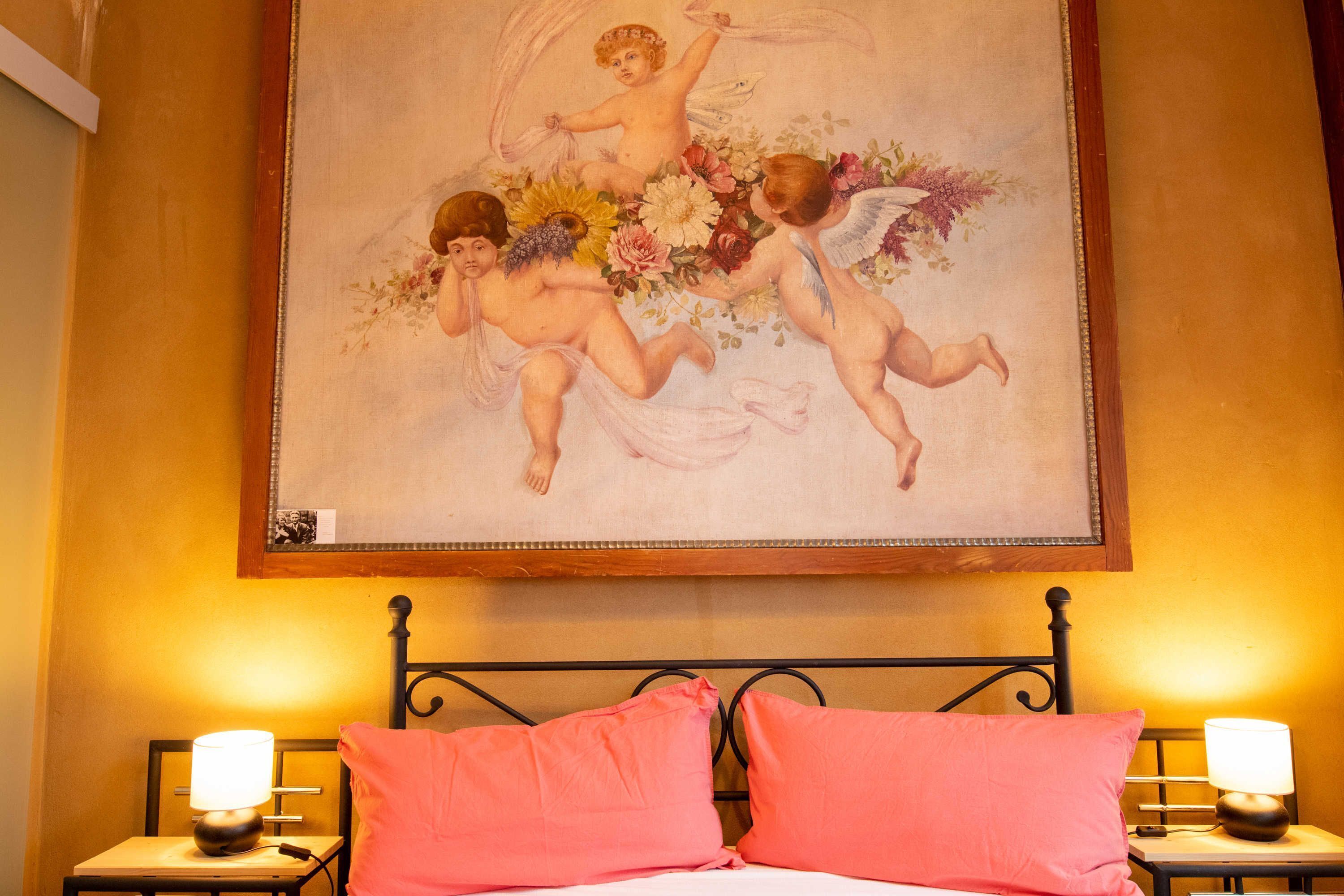 A bedroom with a bed, two bedside tables with lamps, and a painting of cherubs on the wall.