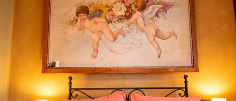 A bedroom with a bed, two bedside tables with lamps, and a painting of cherubs on the wall.