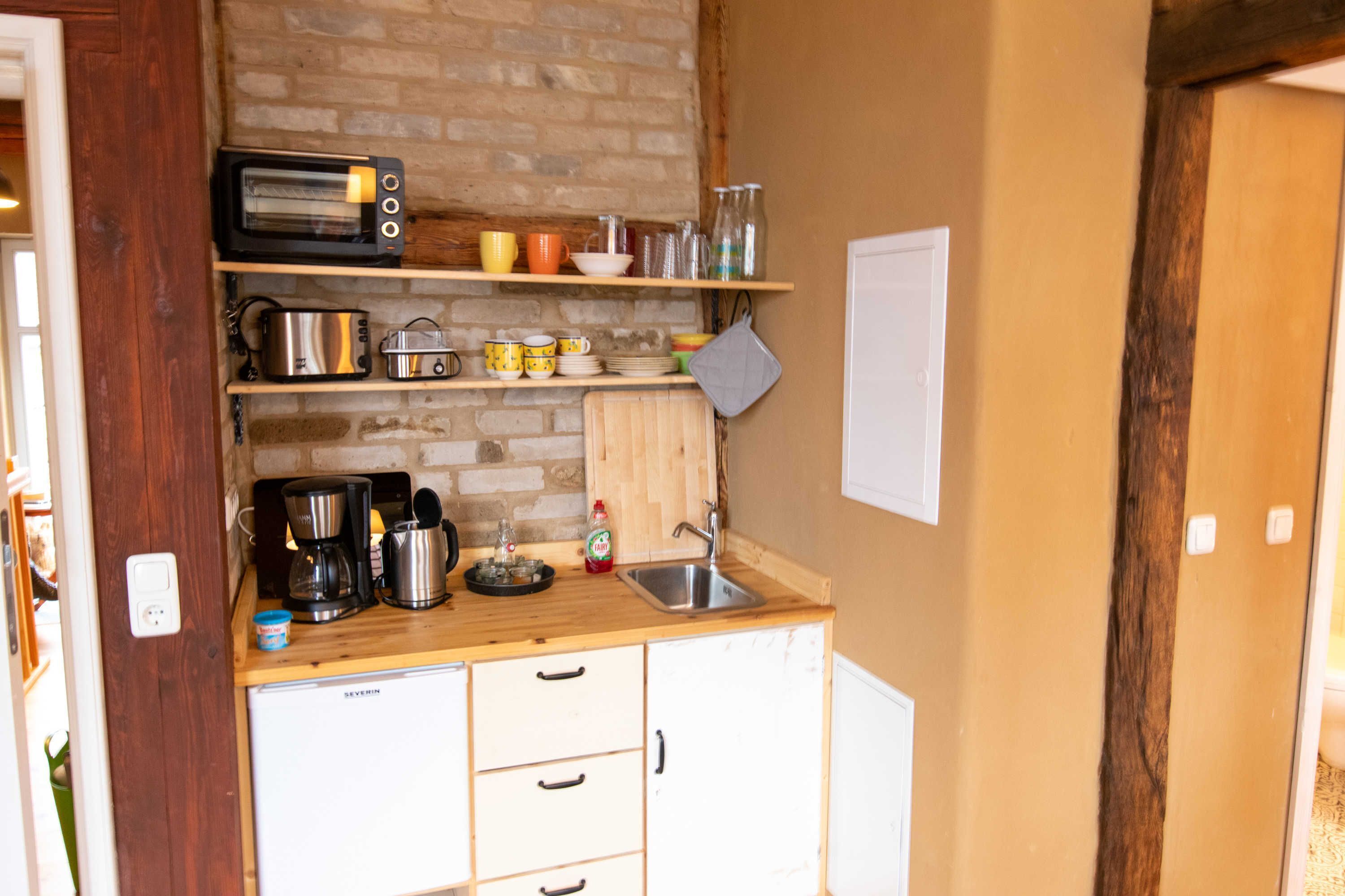 A compact kitchen with a brick wall, wooden shelves, a coffee maker, a toaster, and a sink.