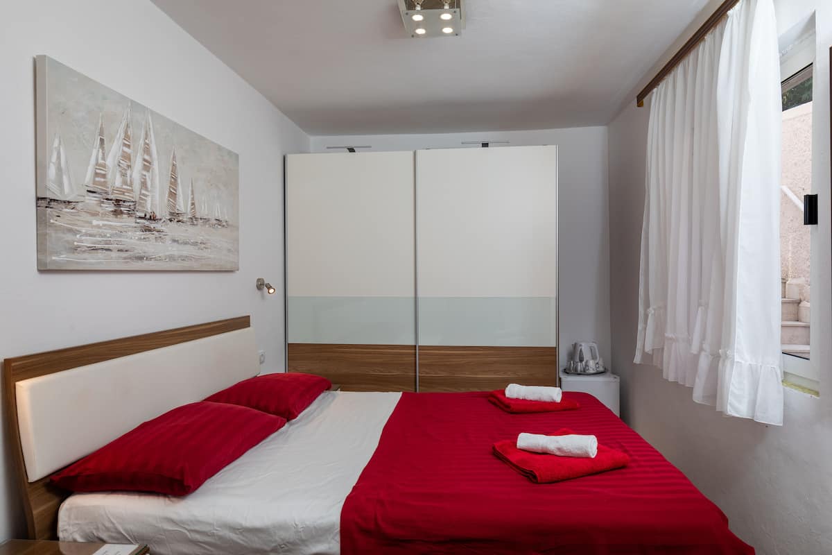 Habitación de hotel con cama, ropa de cama roja, un cuadro en la pared, un armario y una ventana con cortinas.