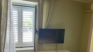 A small room with a desk, a television, a window with curtains, and a trash bin. - Rooms Edna (Dubrovnik)
