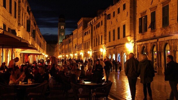 A historic street lined with buildings, featuring outdoor seating areas and a prominent tower in the background. - Rooms Edna (Dubrovnik)