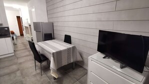 A modern kitchen with white cabinets, a dining table with a patterned tablecloth, two chairs, a TV on a white stand, and a refrigerator. - Renovated Studio - South Balcony - 6 Pers - Ski/Hike (Saint-Michel-de-Chaillol)