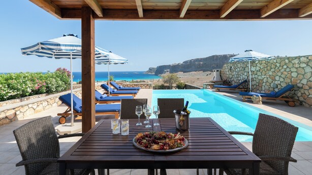 Taula de fusta amb un plat de picar i begudes, amb vistes a una piscina amb gandules i ombrel·les.