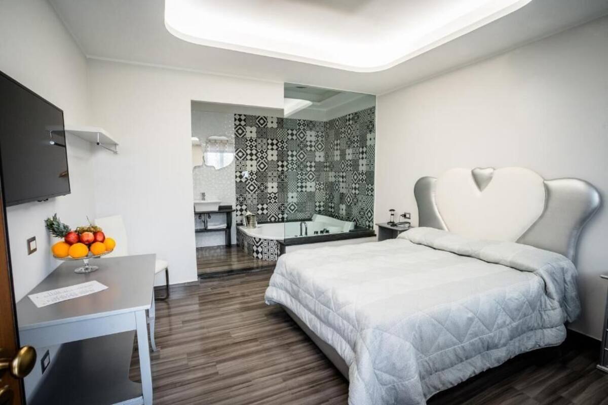 Quarto moderno com cama grande, escrivaninha com frutas, banheiro com azulejos estampados e televisão na parede.