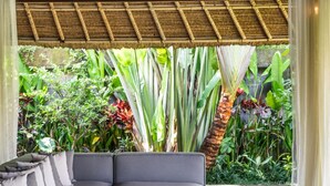 Ein stilvoller Wohnbereich mit einem grauen Ecksofa, einem kleinen runden Holztisch und einem schwarzen Teppich. Der Raum zeichnet sich durch eine strohgedeckte Decke aus und ist von üppigen tropischen Pflanzen umgeben.