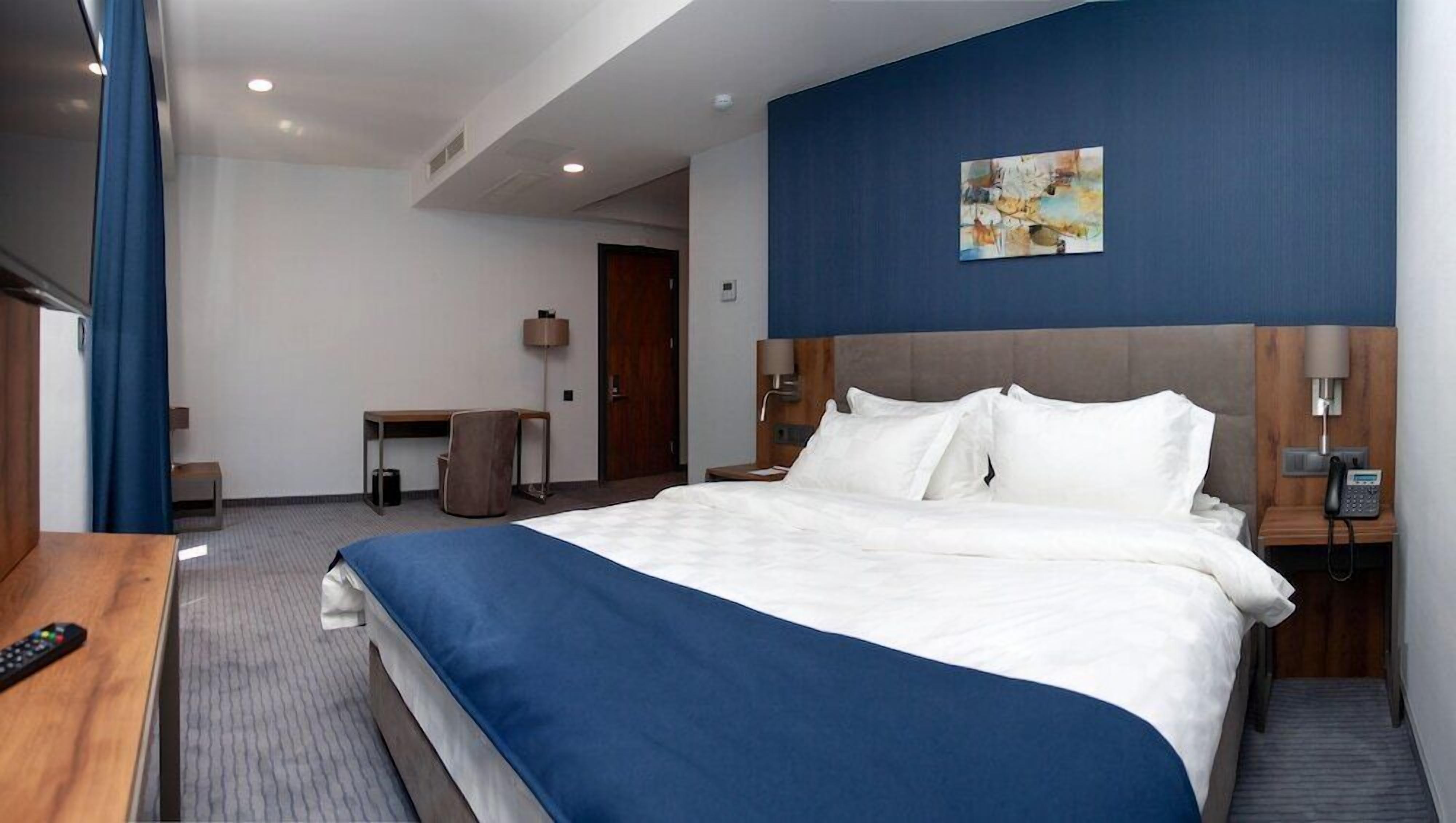A hotel room with a large bed, a desk, a chair, a phone, and a painting on the wall.