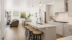 A modern kitchen with white cabinetry, a central island with stools, pendant lighting, and a view into the dining area. - Attitude Adjustment Retreat: Desert Color Lagoon + Beaches (St. George)