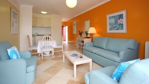 A living room with a blue sofa, a white coffee table, and a dining area with a table and chairs. - Caniço Bay Apartments (Caniço)