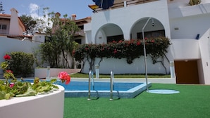 A swimming pool with a blue tiled deck, surrounded by green artificial turf. There are two metal pool ladders, a blue umbrella, and a white wall with a balcony. - Caniço Bay Apartments (Caniço)