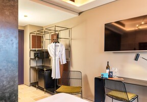 Una habitación de hotel con cama, escritorio con lámpara, televisor montado en la pared, perchero con toallas y silla con cojín.